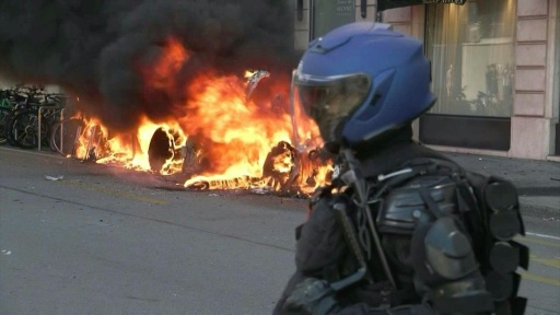 Un véhicule en feu derrière un policier casqué lors de la manifestation anti-G7 à Genève