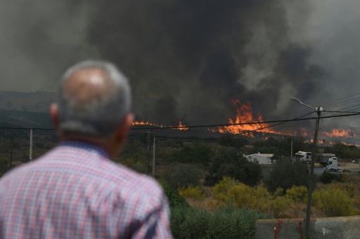 Un homme de dos regarde un front de flammes et une épaisse fumée noire approcher des maisons