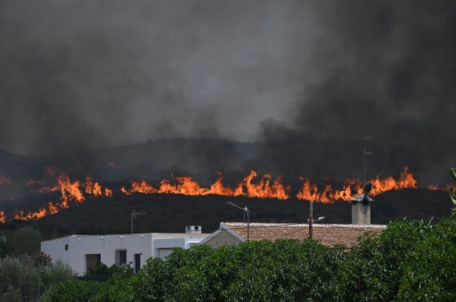 Des flammes vives derrière une maison blanche, sous un ciel noirci par la fumée