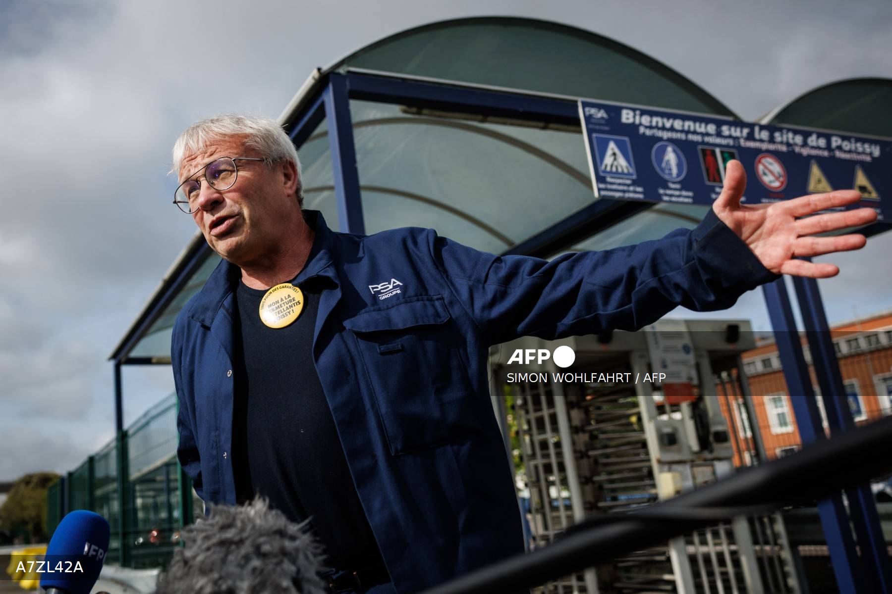 Un syndicaliste en uniforme PSA Groupe, portant un badge «&nbsp;NON À LA FERMETURE DE STELLANTIS POISSY&nbsp;», s'exprime devant l'entrée du site, sous le panneau «&nbsp;Bienvenue sur le site de Poissy&nbsp;»