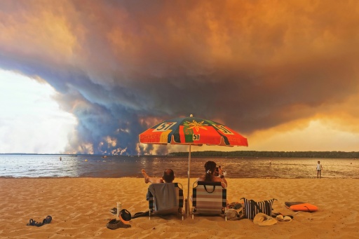 Deux personnes assises sous un parasol sur une plage, face à un immense panache de fumée