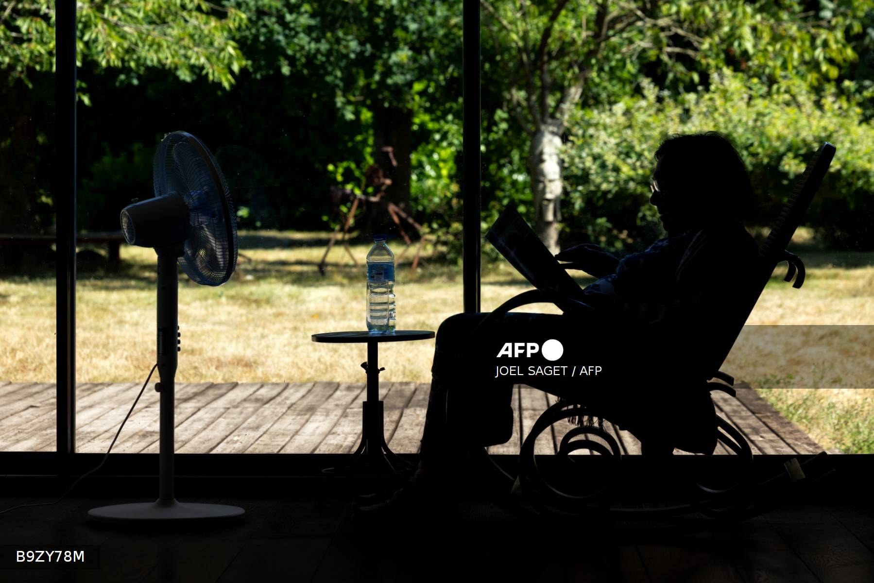 Une personne lit en silhouette devant un ventilateur, face à un jardin desséché, pendant une vague de chaleur